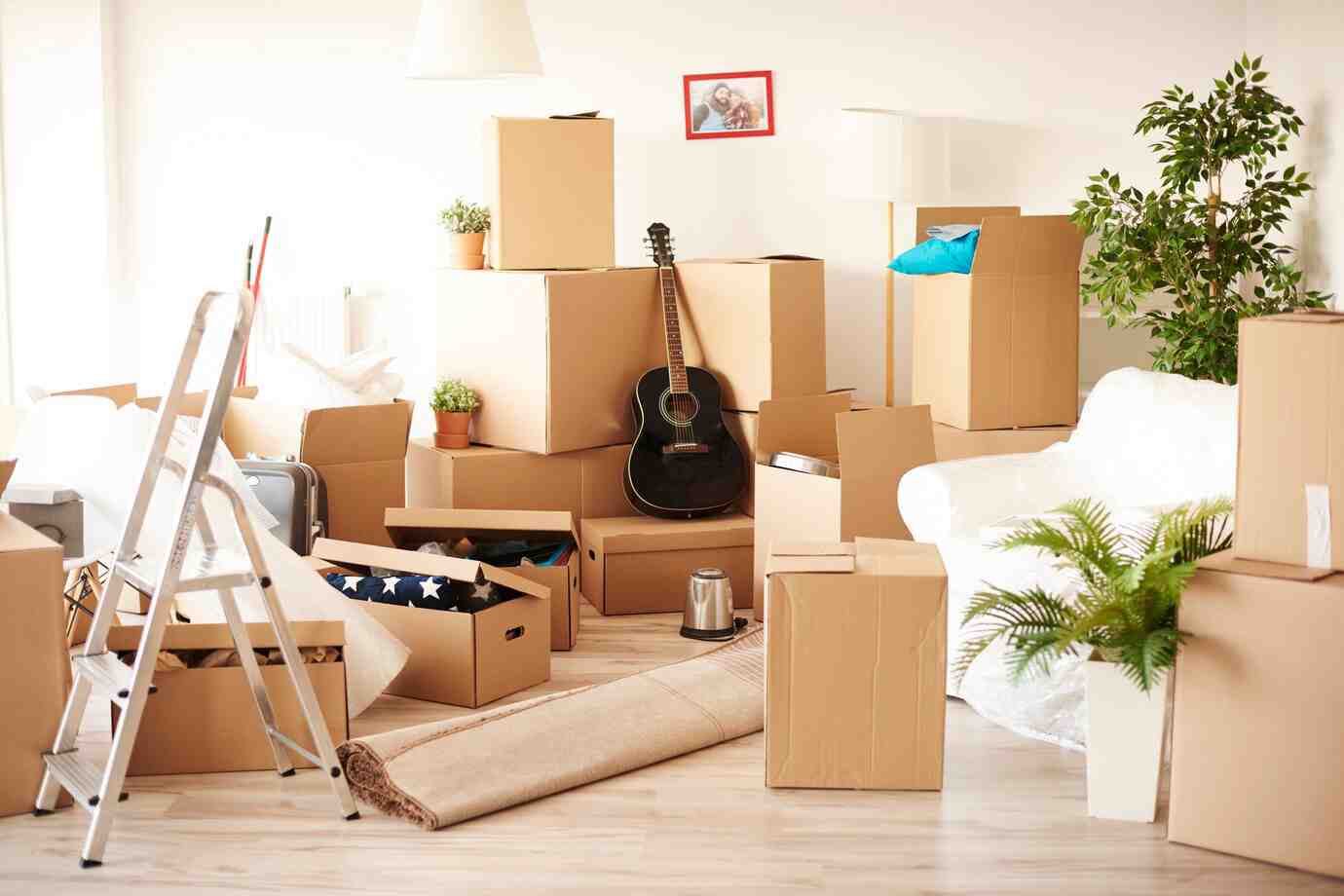 A cluttered room filled with cardboard boxes, a black guitar leaning against them, a ladder, and a white couch partially covered in plastic.