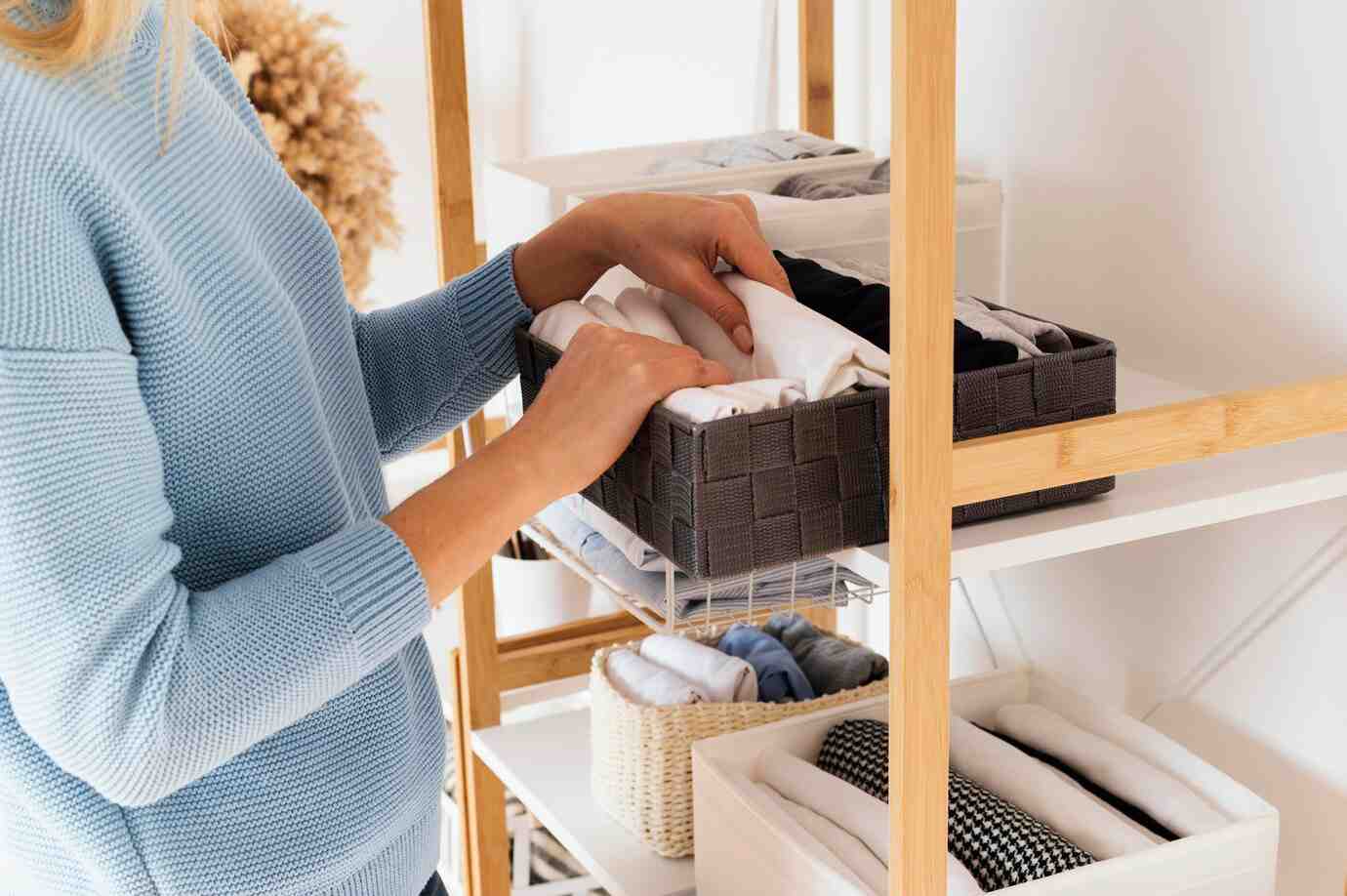 A person in a blue sweater organizes folded clothes in a gray woven basket on a modern wooden shelf.