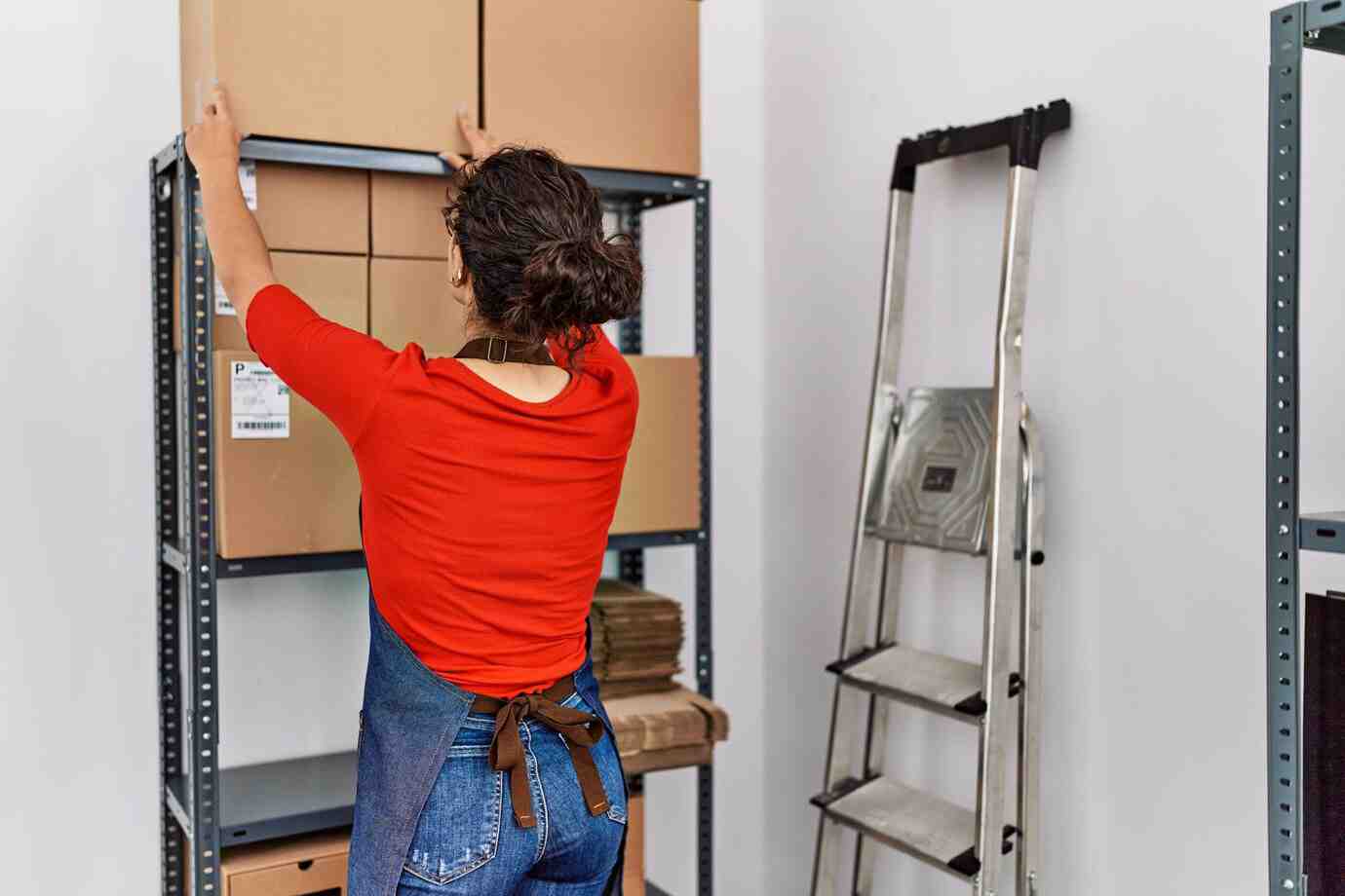 A woman in a red shirt and apron arranges cardboard boxes on a high shelf beside a metal step ladder in a storage area.