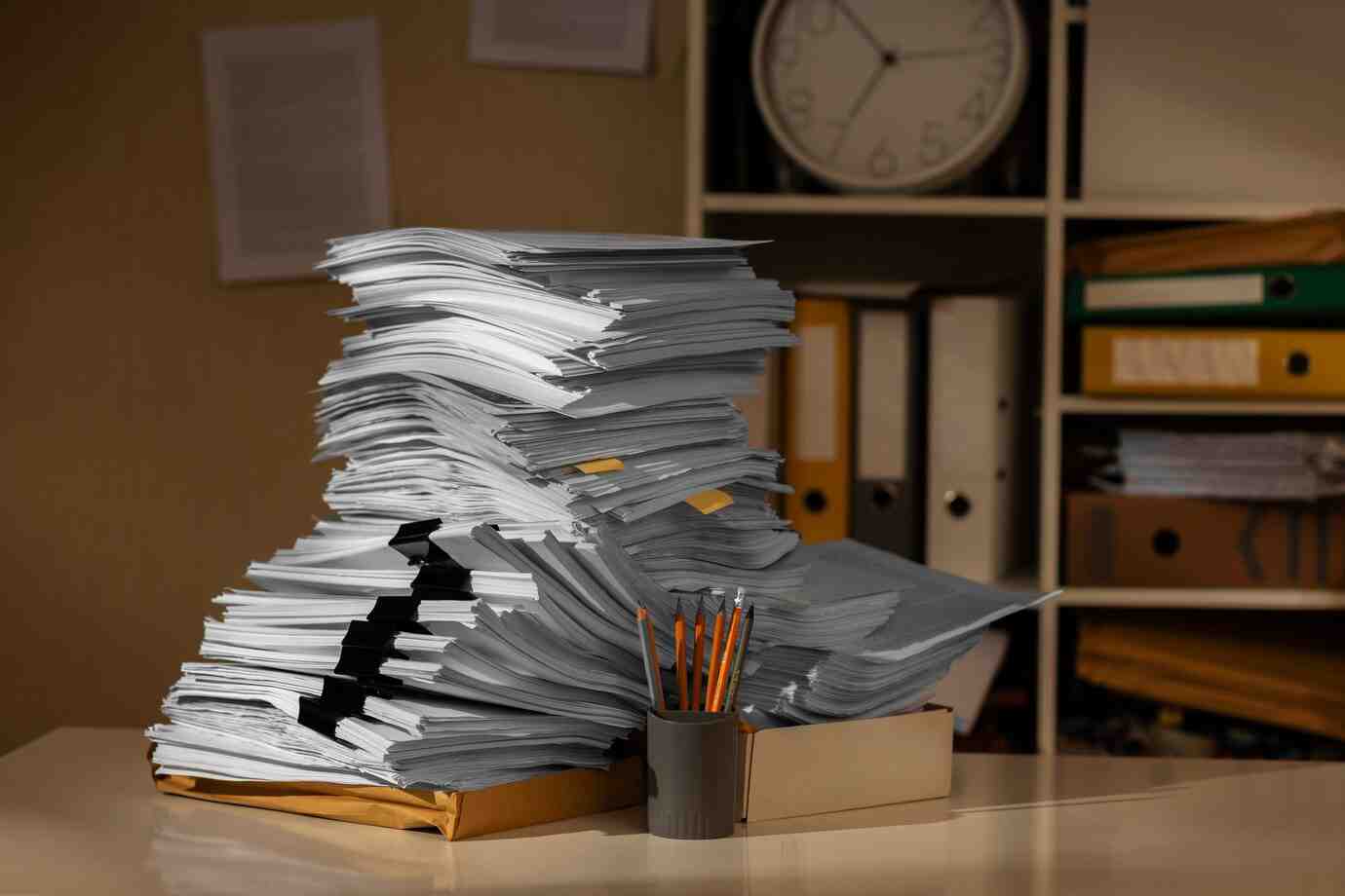 A cluttered workspace featuring a towering stack of papers, a holder with pencils, and organized binders in the background.