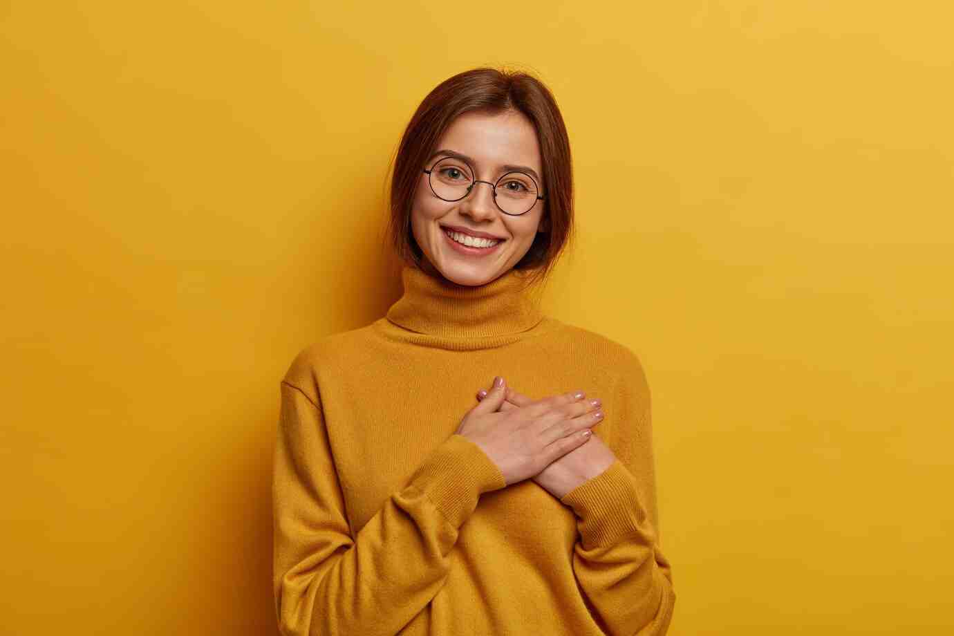 A person wearing a cozy mustard-yellow turtleneck sweater stands against a matching yellow background, hands clasped over their chest.