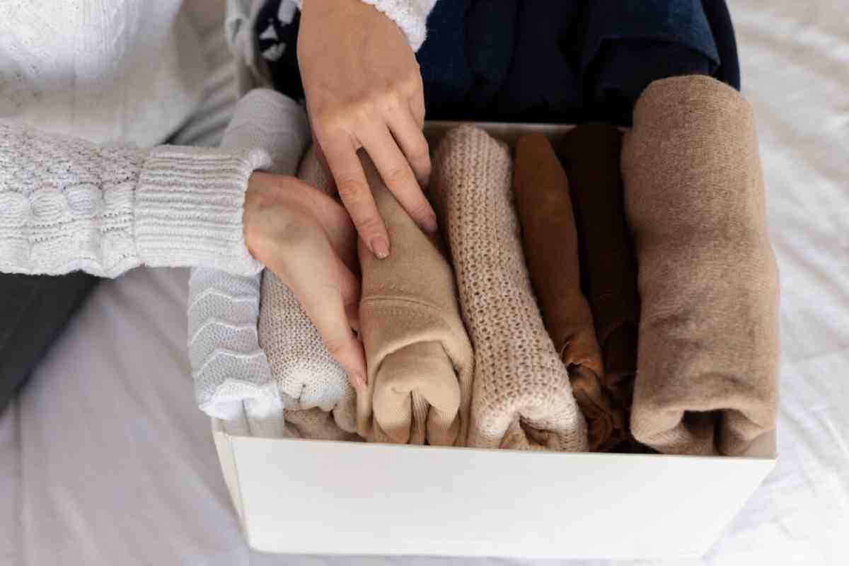 A person arranges neatly rolled clothes in a storage box, placing various shades of fabrics on a bed.