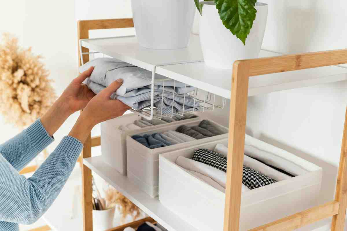 Hands placing neatly folded clothes onto a modern shelf with organized storage bins, surrounded by minimalist decor and plants.
