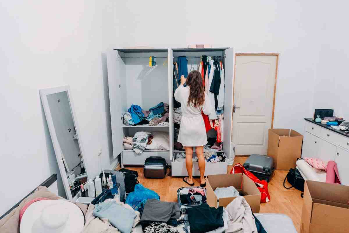 A woman in a robe stands in front of a messy wardrobe, sorting through clothes amidst boxes and a cluttered bedroom.