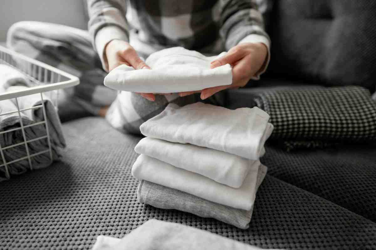 A person in pajamas folds fresh laundry, stacking neatly folded white and gray towels on a textured grey surface. A laundry basket is nearby.