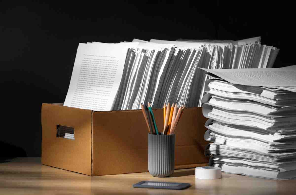 A box overflowing with documents beside a stack of papers, colored pencils in a gray cup, and a small roll of tape on a wooden desk.