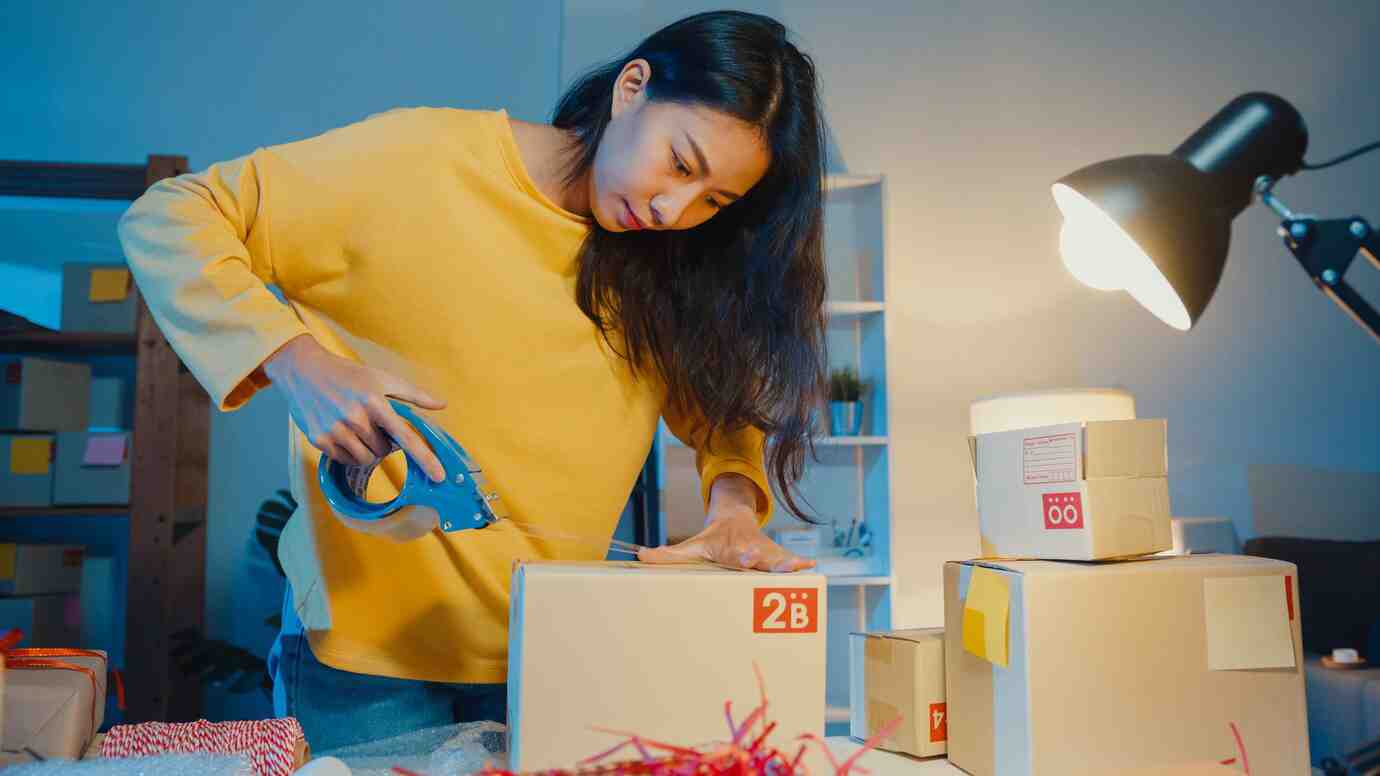 A person in a yellow sweater uses a tape dispenser to seal a box, surrounded by colorful packaging materials and lit by a desk lamp.
