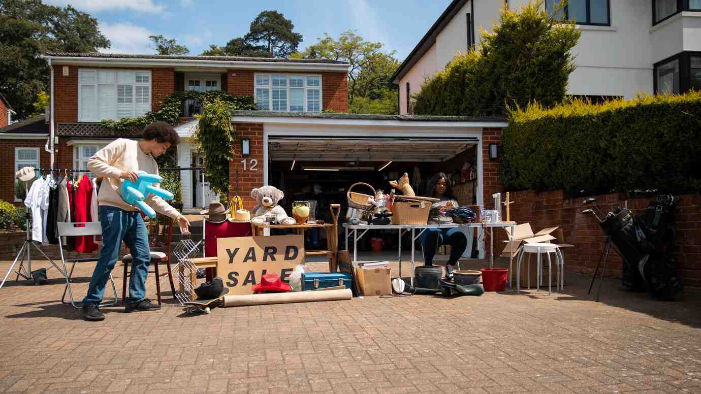 A yard sale displays various items, including clothes, toys, hats, and furniture, outside a suburban home. A sign reads 
