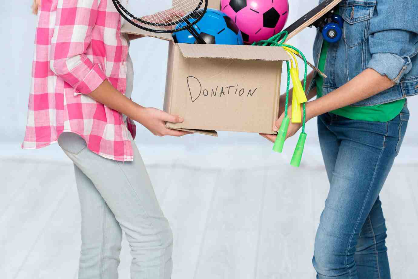 Two children hold a cardboard box labeled DONATION, filled with sports equipment like soccer balls and a tennis racquet.