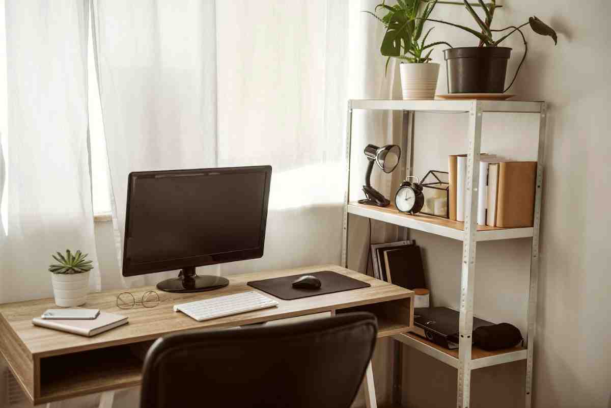 A cozy home office featuring a computer desk, a monitor, plant decor, and a metal shelf with books and a desk lamp. Natural light filters through curtains.