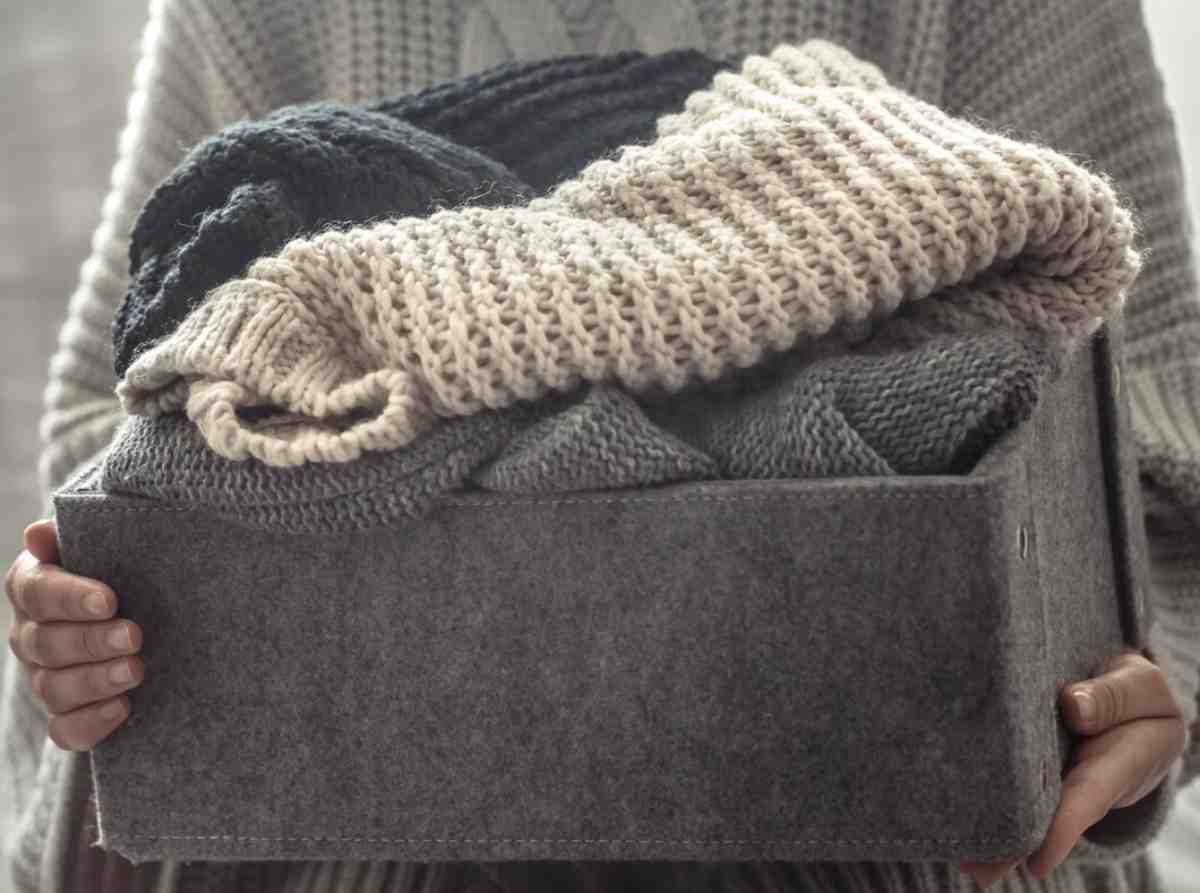 A person holds a gray felt box filled with cozy knitted sweaters in various neutral colors. Soft textiles create a warm, inviting look.