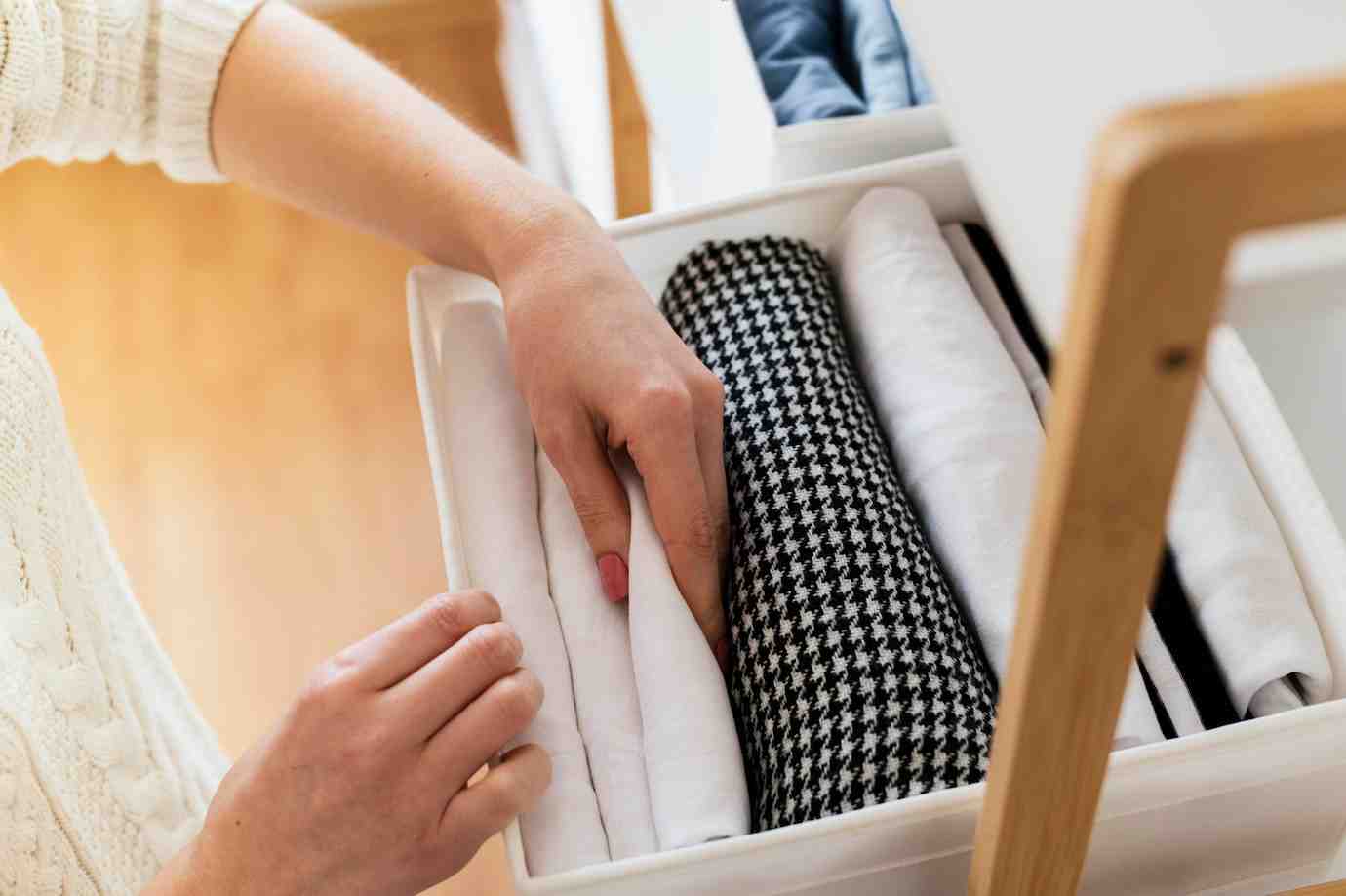 A person with a knitted sweater neatly organizes rolled clothes in a drawer, featuring white, black, and patterned fabrics.