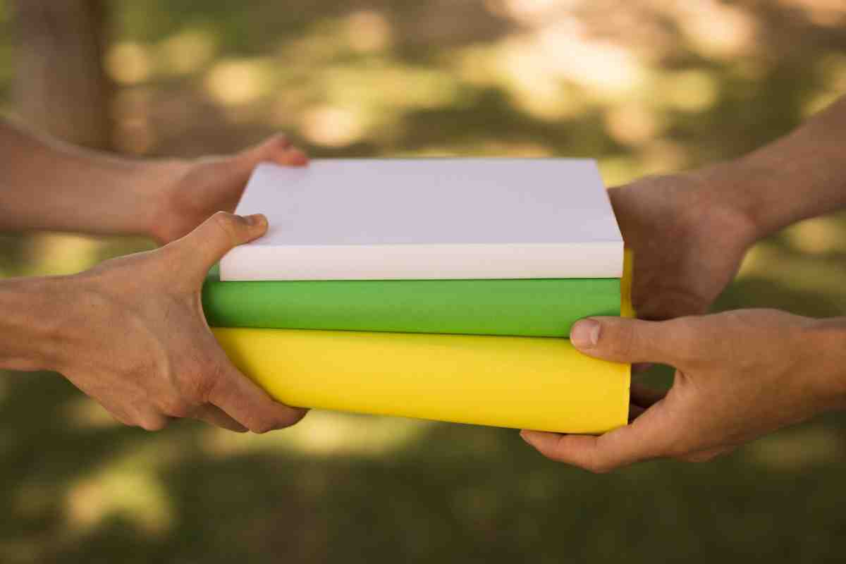 Two hands exchanging a stack of vibrant, colorful books—white, green, and yellow—against a softly blurred outdoor background.