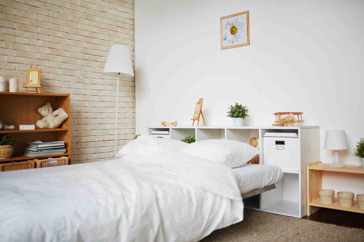 Cozy bedroom featuring a white bed, teddy bears, plants, and light wood furniture against a brick wall. Warm, inviting atmosphere.