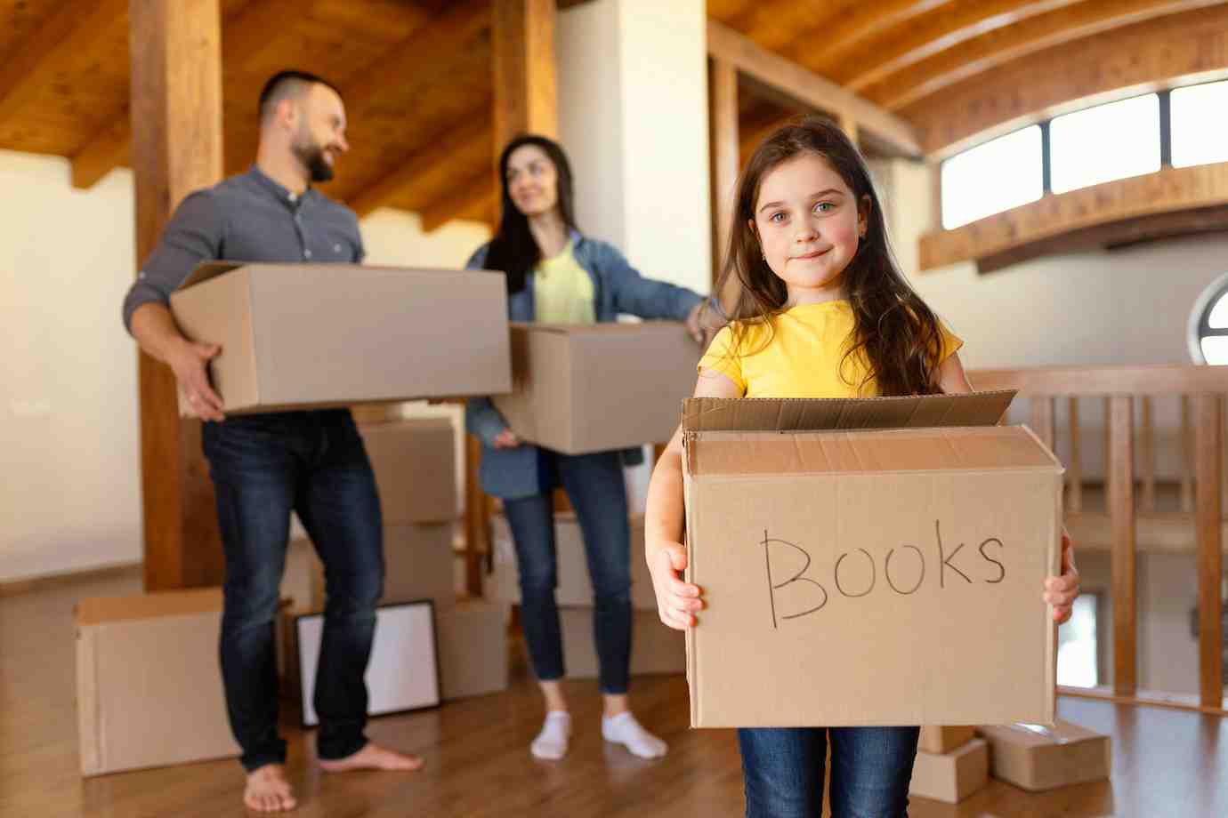 A girl in a yellow shirt holds a cardboard box labeled Books, while two adults carry larger boxes in a bright, spacious room.