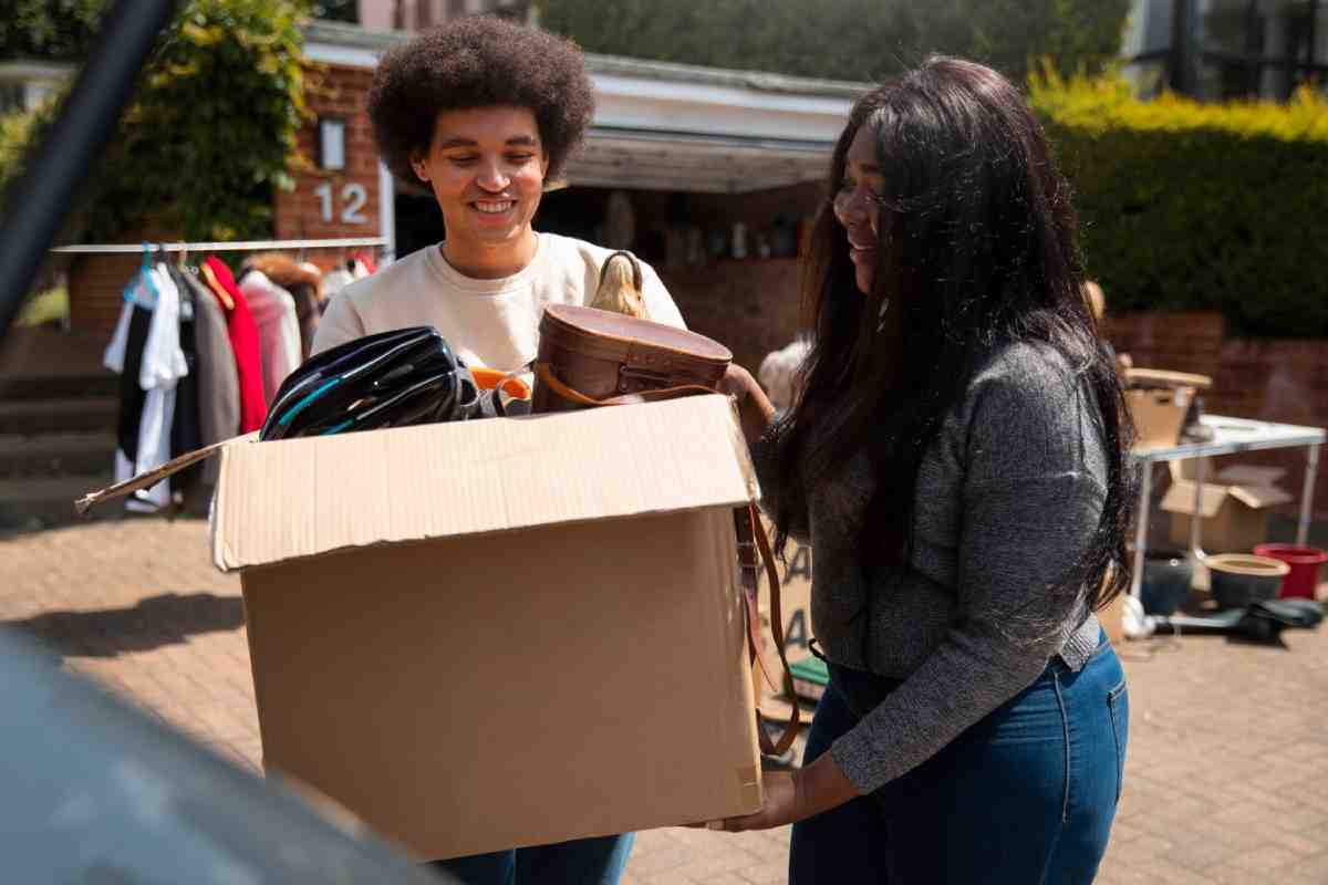 Two individuals are carrying a box filled with clothing and bags outside during a sunny day, with more items displayed in the background.