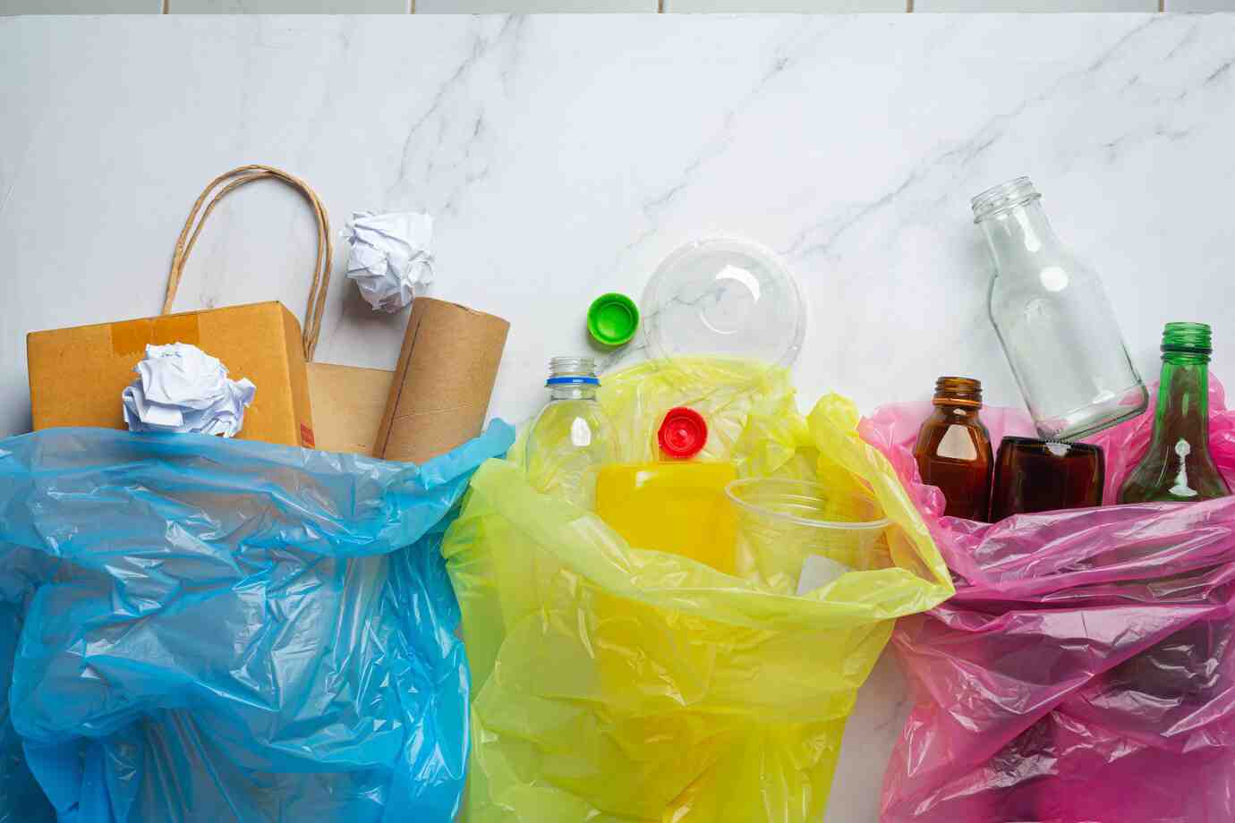 Three colored garbage bags on a marble surface: blue with paper, yellow with plastic, and pink with glass bottles and jars.