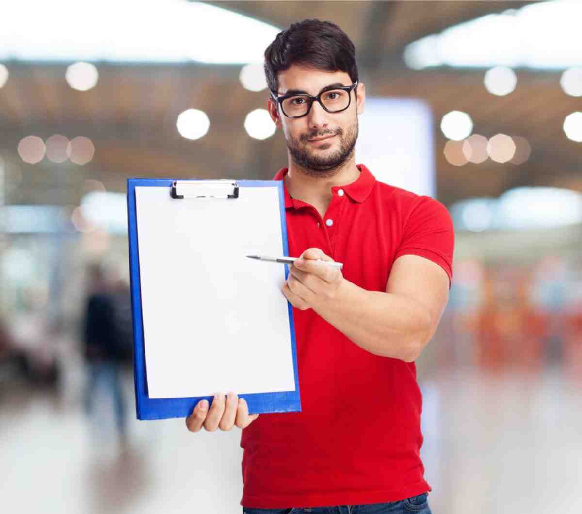 Person in a red shirt holds a clipboard with a blank sheet, standing in a bright, blurred indoor setting.