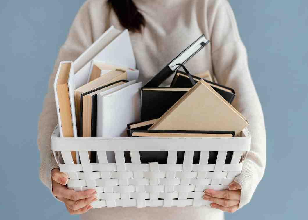 A person holds a white woven basket filled with assorted books, both blank and covered, against a soft blue background.