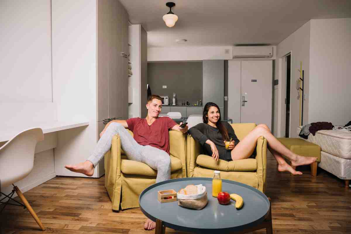 A cozy living room scene featuring two individuals relaxing on yellow sofas with a coffee table displaying snacks and drinks.