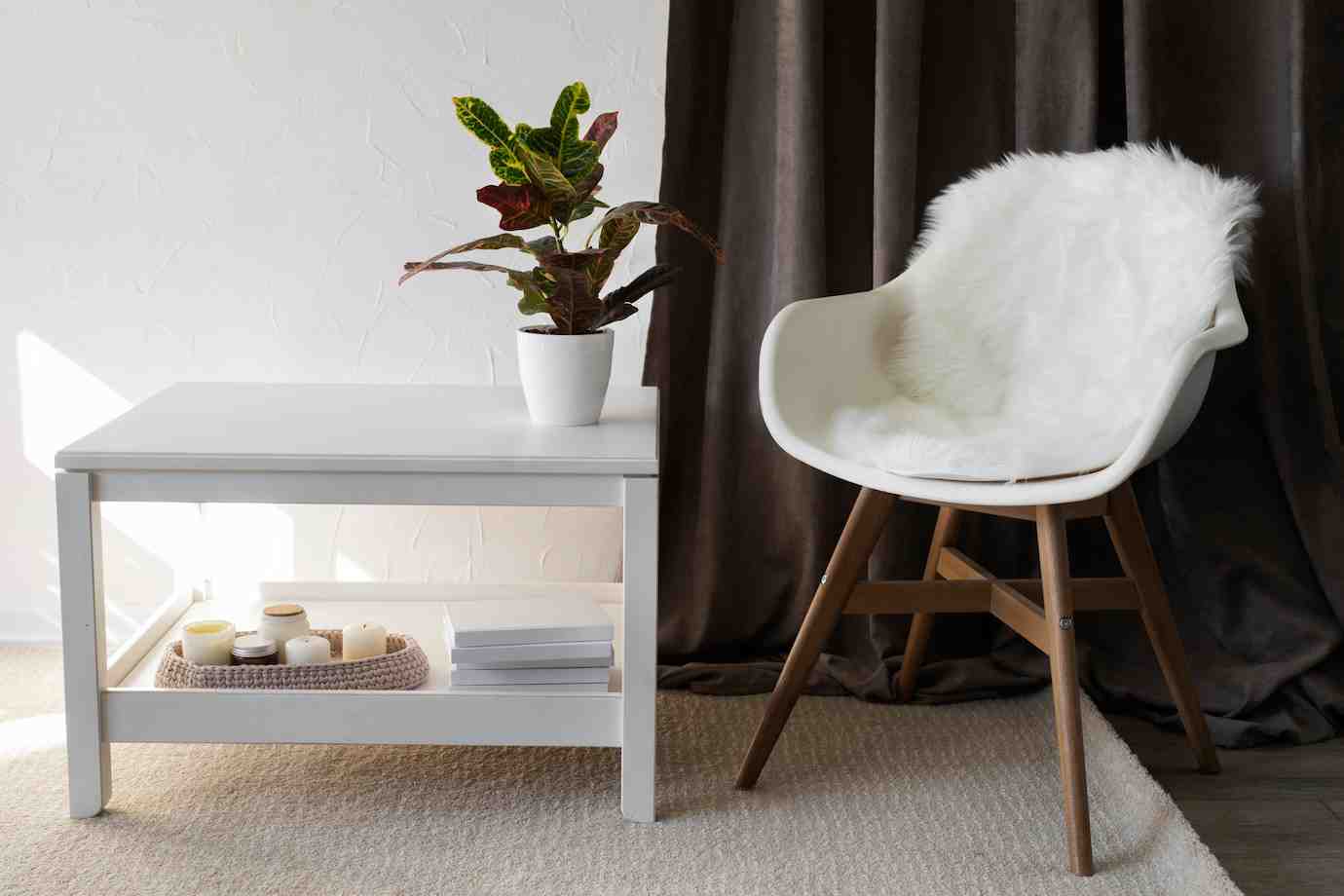 A white coffee table with a potted plant and candles beside a cozy chair draped in white fur, against a backdrop of dark curtains.