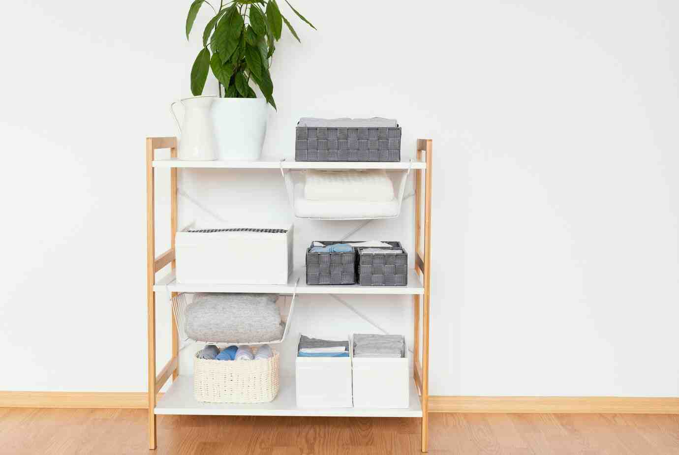A stylish white shelf with wooden accents, featuring neatly organized storage boxes, towels, and a potted plant on top.